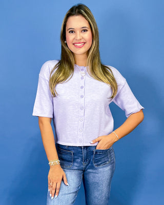 A woman modeling a light lavender short-sleeve button-up knit top with blue jeans, standing against a solid blue studio background.