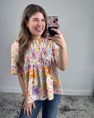 Smiling woman taking a mirror selfie wearing a colorful floral short-sleeve blouse with a pleated front, paired with dark-wash straight-leg jeans and white pointed-toe heels in a neutral studio setting.