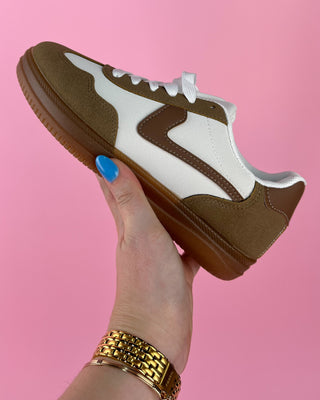 Brown and white sneaker held by a hand with blue nail polish against a pink background