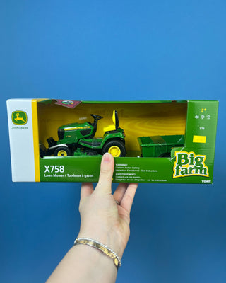 A hand holds a boxed John Deere toy lawn mower and trailer set against a solid blue background. The packaging shows a green and yellow X758 lawn mower with a green trailer, and the boxes include John Deere and Big Farm branding.