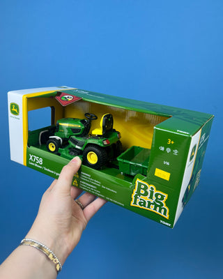A hand holds a boxed John Deere toy lawn mower and trailer set against a solid blue background. The packaging shows a green and yellow X758 lawn mower with a green trailer, and the boxes include John Deere and Big Farm branding.