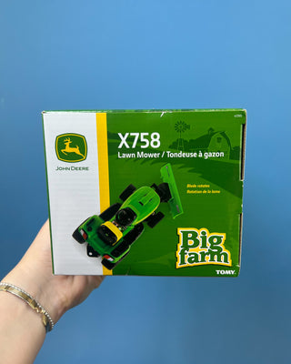 A hand holds a boxed John Deere toy lawn mower and trailer set against a solid blue background. The packaging shows a green and yellow X758 lawn mower with a green trailer, and the boxes include John Deere and Big Farm branding.