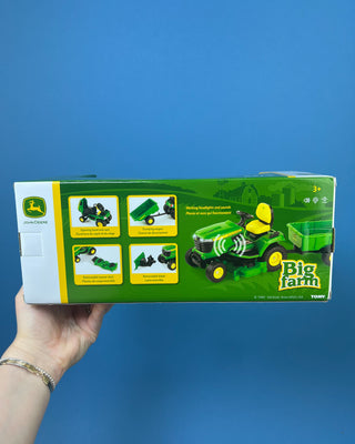 A hand holds a boxed John Deere toy lawn mower and trailer set against a solid blue background. The packaging shows a green and yellow X758 lawn mower with a green trailer, and the boxes include John Deere and Big Farm branding.