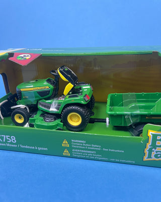 A hand holds a boxed John Deere toy lawn mower and trailer set against a solid blue background. The packaging shows a green and yellow X758 lawn mower with a green trailer, and the boxes include John Deere and Big Farm branding.