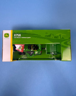 A hand holds a boxed John Deere toy lawn mower and trailer set against a solid blue background. The packaging shows a green and yellow X758 lawn mower with a green trailer, and the boxes include John Deere and Big Farm branding.
