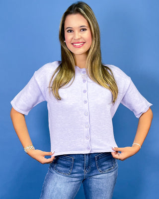A woman with blonde-highlighted hair wearing a light lavender short-sleeve button-up top and blue jeans, posing against a solid blue studio background.