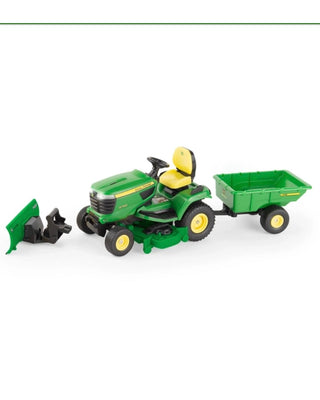 A hand holds a boxed John Deere toy lawn mower and trailer set against a solid blue background. The packaging shows a green and yellow X758 lawn mower with a green trailer, and the boxes include John Deere and Big Farm branding.