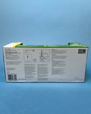 A hand holds a boxed John Deere toy lawn mower and trailer set against a solid blue background. The packaging shows a green and yellow X758 lawn mower with a green trailer, and the boxes include John Deere and Big Farm branding.
