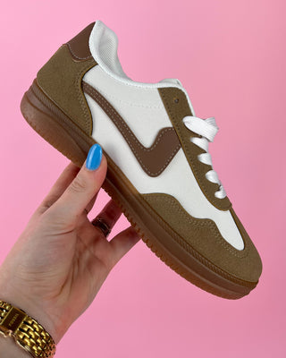 Brown and white sneaker held by a hand with blue nail polish against a pink background