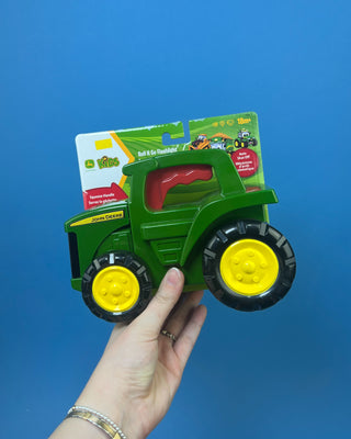 A green John Deere toy tractor held in a person's hand against a solid blue background. The toy has yellow wheels and a red seat, and it is still attached to its original retail packaging card.