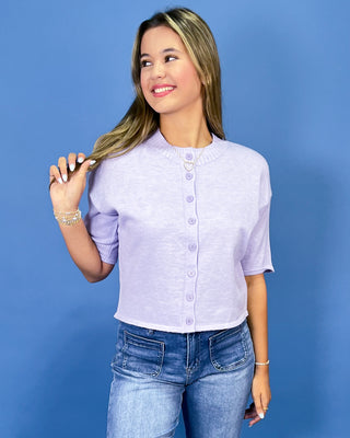a light lavender short-sleeve button-up top and blue jeans, posing against a solid blue studio background.