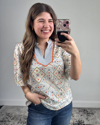 Smiling woman taking a mirror selfie wearing a short-sleeve patterned blouse with a striped split neckline and orange trim, paired with dark-wash straight-leg jeans and white pointed-toe heels in a neutral studio setting.