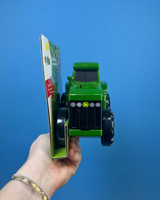 A green John Deere toy tractor held in a person's hand against a solid blue background. The toy has yellow wheels and a red seat, and it is still attached to its original retail packaging card.