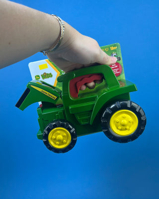 A green John Deere toy tractor held in a person's hand against a solid blue background. The toy has yellow wheels and a red seat, and it is still attached to its original retail packaging card.