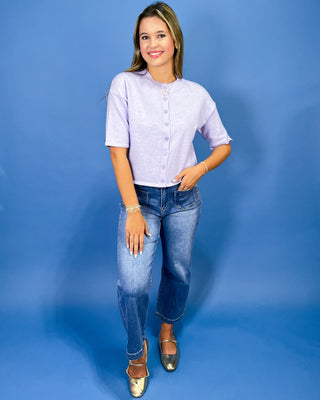 A woman modeling a light lavender short-sleeve button-up top with blue jeans and metallic gold shoes, standing against a solid blue studio background.