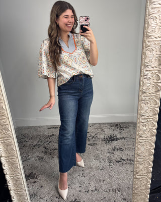 Smiling woman taking a mirror selfie wearing a short-sleeve patterned blouse with a striped split neckline and orange trim, paired with dark-wash straight-leg jeans and white pointed-toe heels in a neutral studio setting.