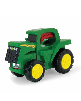A green John Deere toy tractor held in a person's hand against a solid blue background. The toy has yellow wheels and a red seat, and it is still attached to its original retail packaging card.