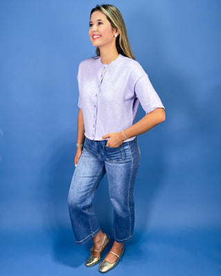A woman modeling a light lavender short-sleeve button-up top with blue jeans and metallic gold flats, standing against a solid blue studio backdrop.