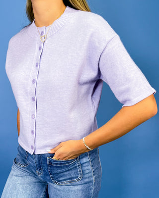 A woman wearing a light lavender short-sleeve knit cardigan with a button-up front and a small gold ring detail at the collar, paired with blue jeans against a solid blue background.