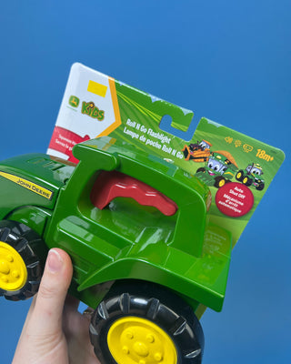 A green John Deere toy tractor held in a person's hand against a solid blue background. The toy has yellow wheels and a red seat, and it is still attached to its original retail packaging card.