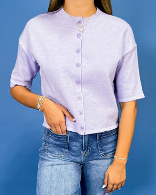 a light lavender short-sleeve button-up top and blue jeans, posing against a solid blue studio background.