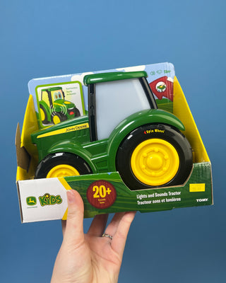 A hand holds a boxed John Deere toy tractor for kids. The package contains a green and yellow toy tractor with black wheels, and the box is branded with the John Deere logo and a '20+' sounds badge.