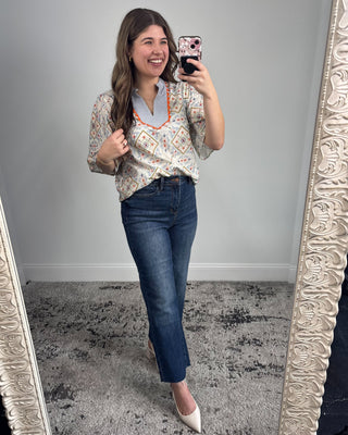 Smiling woman taking a mirror selfie wearing a short-sleeve patterned blouse with a striped split neckline and orange trim, paired with dark-wash straight-leg jeans and white pointed-toe heels in a neutral studio setting.