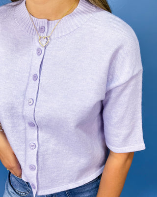 A close-up of a person wearing a light lavender ribbed knit short-sleeve cardigan with matching buttons, paired with blue jeans and a heart-shaped necklace, set against a solid blue background.