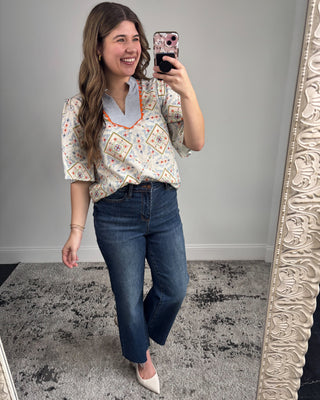 Smiling woman taking a mirror selfie wearing a short-sleeve patterned blouse with a striped split neckline and orange trim, paired with dark-wash straight-leg jeans and white pointed-toe heels in a neutral studio setting.