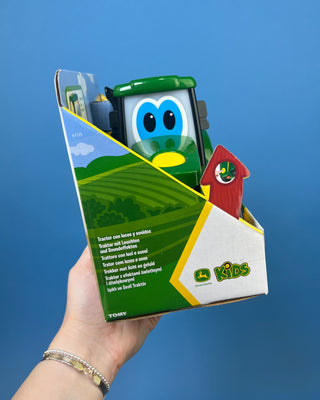 A hand holds a boxed John Deere toy tractor for kids. The package contains a green and yellow toy tractor with black wheels, and the box is branded with the John Deere logo and a '20+' sounds badge.