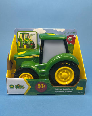A hand holds a boxed John Deere toy tractor for kids. The package contains a green and yellow toy tractor with black wheels, and the box is branded with the John Deere logo and a '20+' sounds badge.