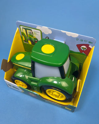 A hand holds a boxed John Deere toy tractor for kids. The package contains a green and yellow toy tractor with black wheels, and the box is branded with the John Deere logo and a '20+' sounds badge.