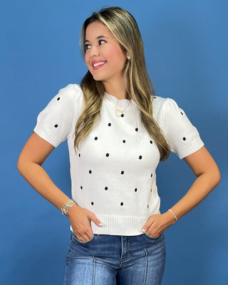 A woman wearing a white short-sleeve knit top with black polka dots, paired with blue jeans, standing against a solid blue studio background.