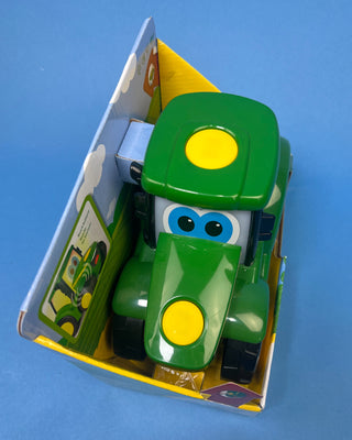 A hand holds a boxed John Deere toy tractor for kids. The package contains a green and yellow toy tractor with black wheels, and the box is branded with the John Deere logo and a '20+' sounds badge.
