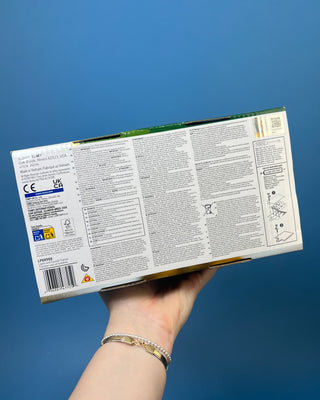 A hand holds a boxed John Deere toy tractor for kids. The package contains a green and yellow toy tractor with black wheels, and the box is branded with the John Deere logo and a '20+' sounds badge.