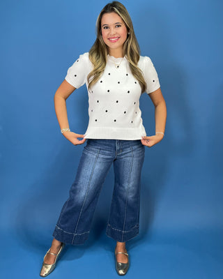 g a short-sleeve cream knit sweater with black polka dots, paired with blue jeans, against a solid blue studio background.