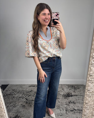 Smiling woman taking a mirror selfie wearing a short-sleeve patterned blouse with a striped split neckline and orange trim, paired with dark-wash straight-leg jeans and white pointed-toe heels in a neutral studio setting.
