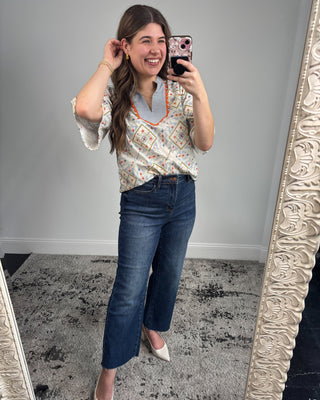 Smiling woman taking a mirror selfie wearing a short-sleeve patterned blouse with a striped split neckline and orange trim, paired with dark-wash straight-leg jeans and white pointed-toe heels in a neutral studio setting.