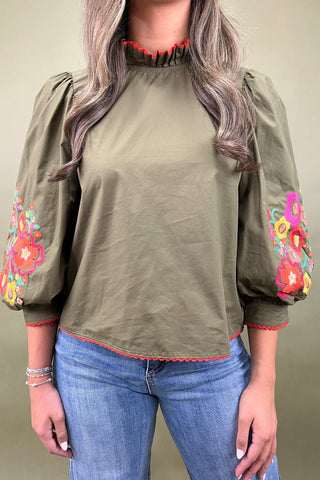 Woman wearing an olive green blouse with colorful floral embroidery on the sleeves against a neutral background