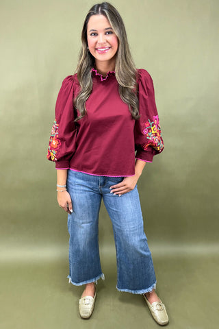 Woman wearing a burgundy blouse with floral embroidery and blue jeans on a beige background