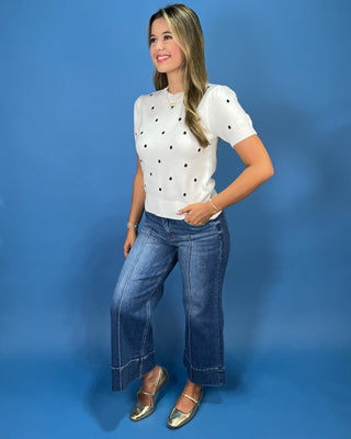 A woman modeling a white short-sleeve knit top with black polka dots, paired with blue wide-leg jeans and metallic gold flats, set against a solid blue studio backdrop.