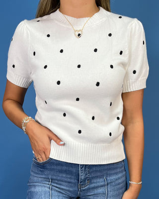 a short-sleeve cream knit sweater with black polka dots, paired with blue jeans, against a solid blue studio background.