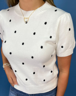 A woman wearing a white short-sleeve knit sweater with black polka dots, paired with blue jeans and a gold necklace, set against a solid blue background.