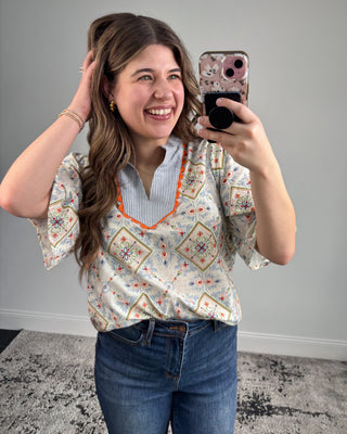 Smiling woman taking a mirror selfie wearing a short-sleeve patterned blouse with a striped split neckline and orange trim, paired with dark-wash straight-leg jeans and white pointed-toe heels in a neutral studio setting.