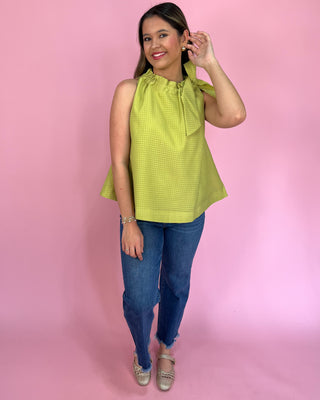 Woman wearing a green sleeveless top with a bow detail and blue jeans against a pink background