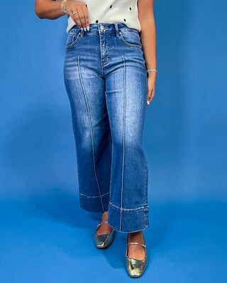 A person wearing blue wide-leg cropped jeans with contrast stitching, paired with a white top and metallic silver flats, standing against a solid blue background.