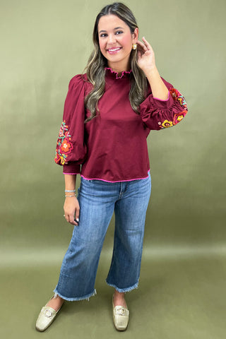 Woman wearing a burgundy blouse with floral embroidery and blue jeans on a green background