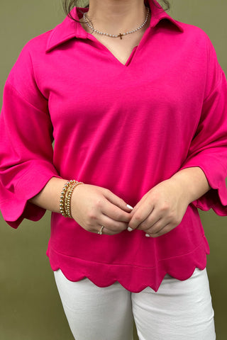 Person wearing a bright pink scalloped blouse with white pants against a green background