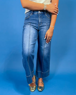 A person wearing medium-wash blue wide-leg cropped jeans with rolled cuffs, paired with metallic gold flats, standing against a solid blue studio background.