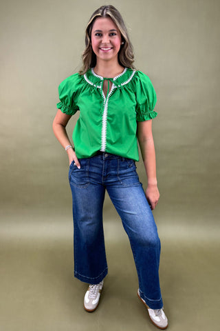 Woman wearing a green blouse with white trim and blue jeans against a beige background